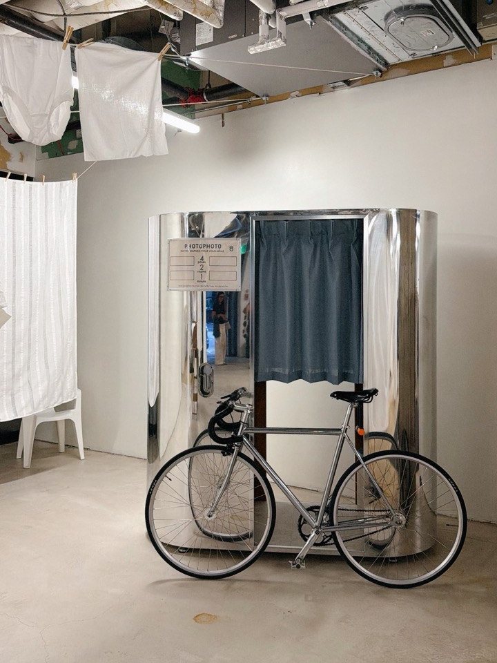 A mirror-polished PHOTOPHOTO booth in a white room, a bicycle leaning against it
